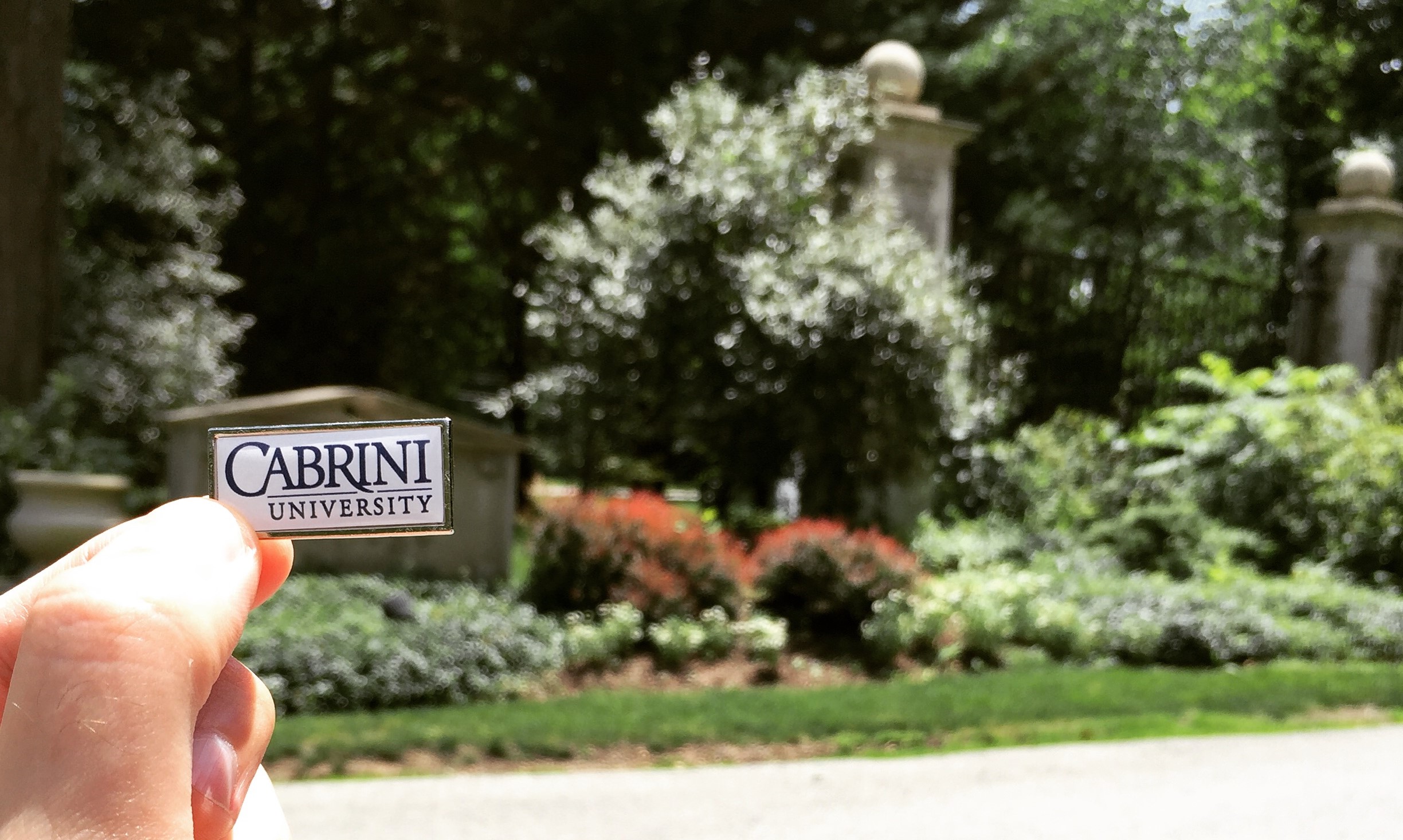 A hand holding a Cabrini University pin over the Cabrini College sign